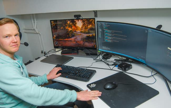 En man sitter med hörlurar vid ett skrivbord med två bildskärmar varav en är en panoramaskärm.
