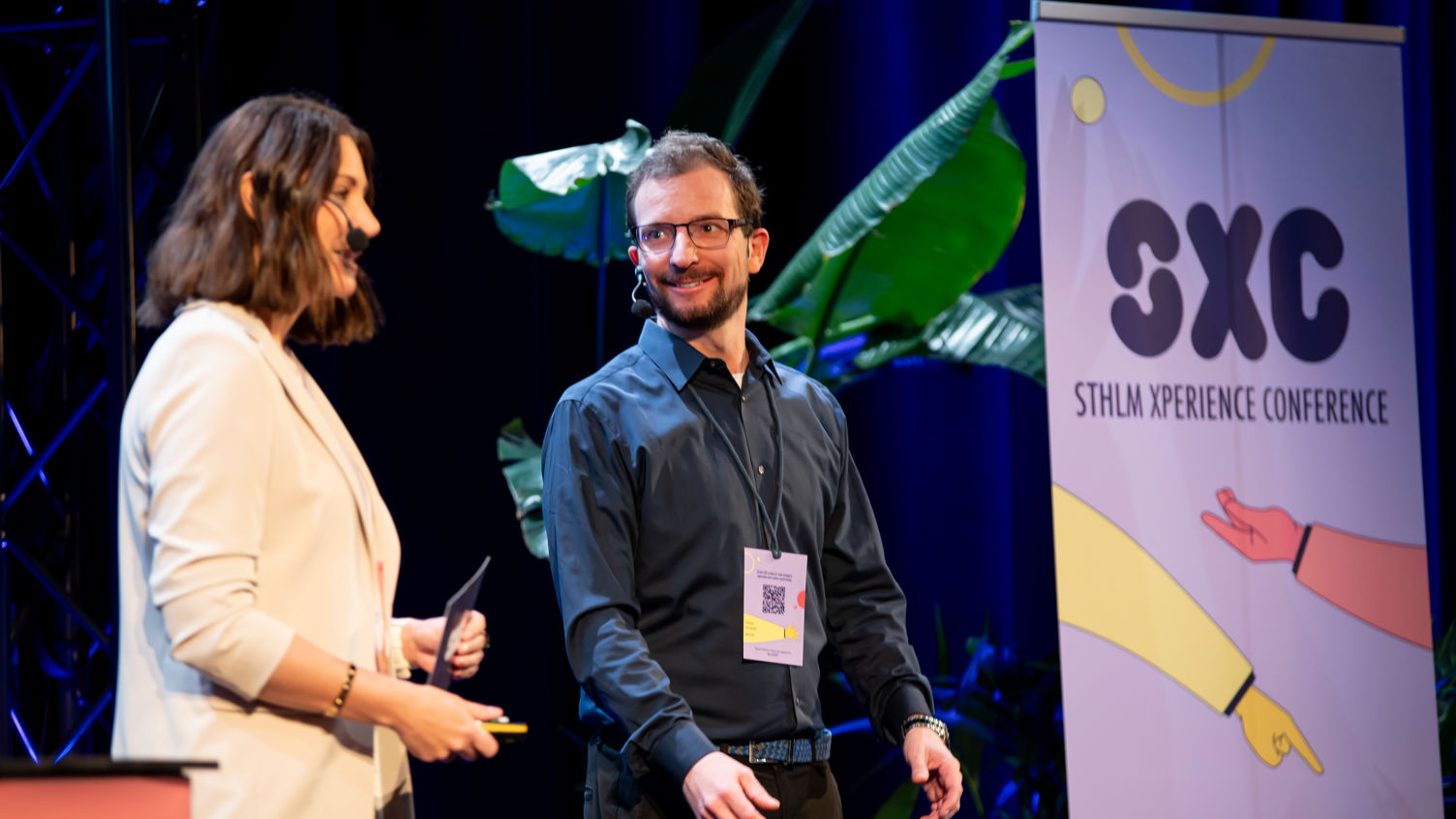 En kvinna och en man står bredvid varandra på en scen med en roll-up i bakgrunden med texten STHLM Xperience Conference