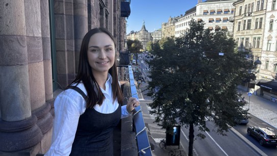 Dana Leshchuk iklädd vit skjorta står leende på Zingtons balkong med sol i bakgrunden.