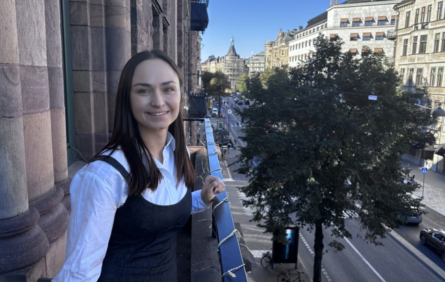 Dana Leshchuk iklädd vit skjorta står leende på Zingtons balkong med sol i bakgrunden.