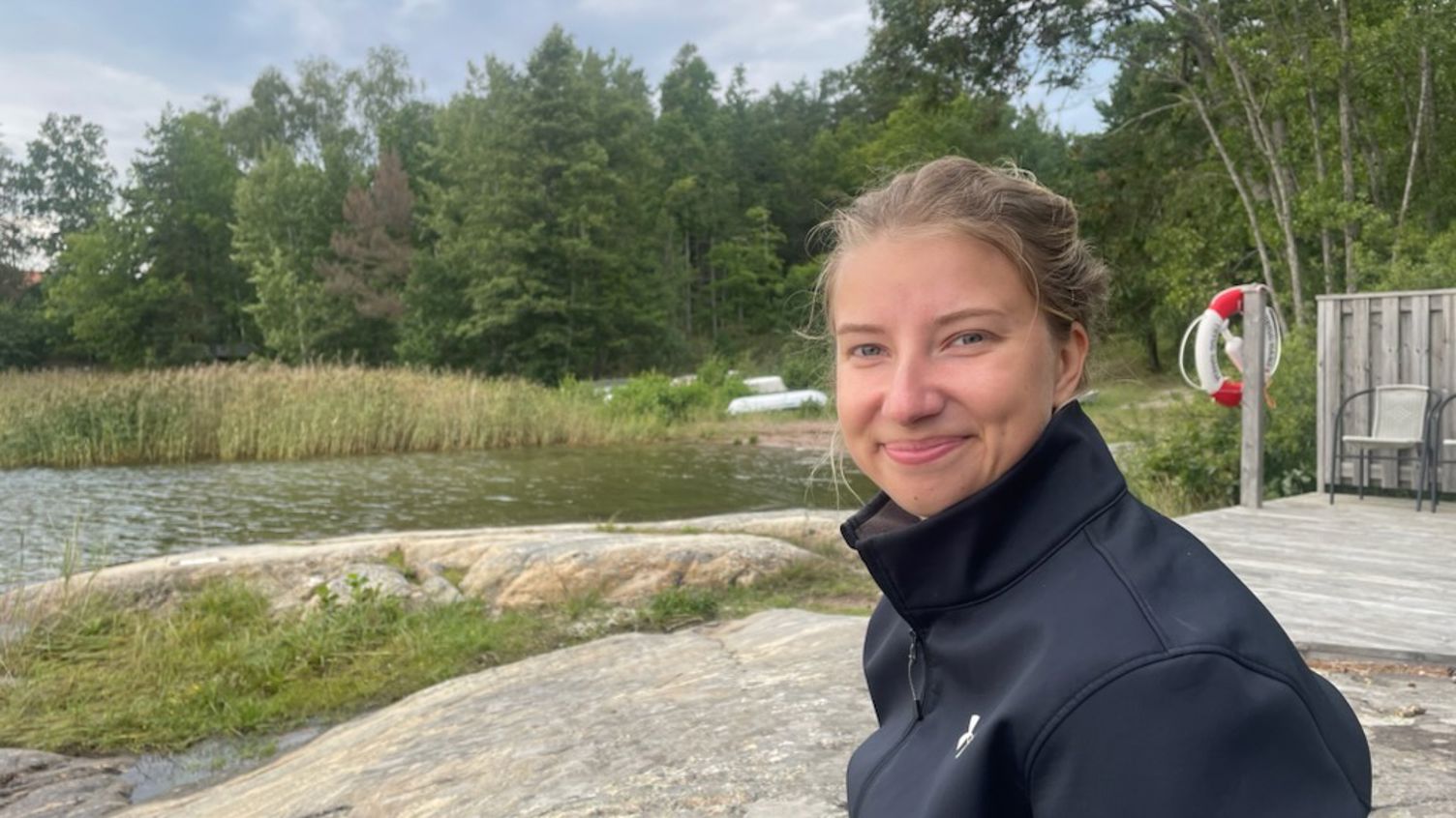 Emelie Djenfer sitter på en klippa och ler, vatten och vass syns i bakgrunden.