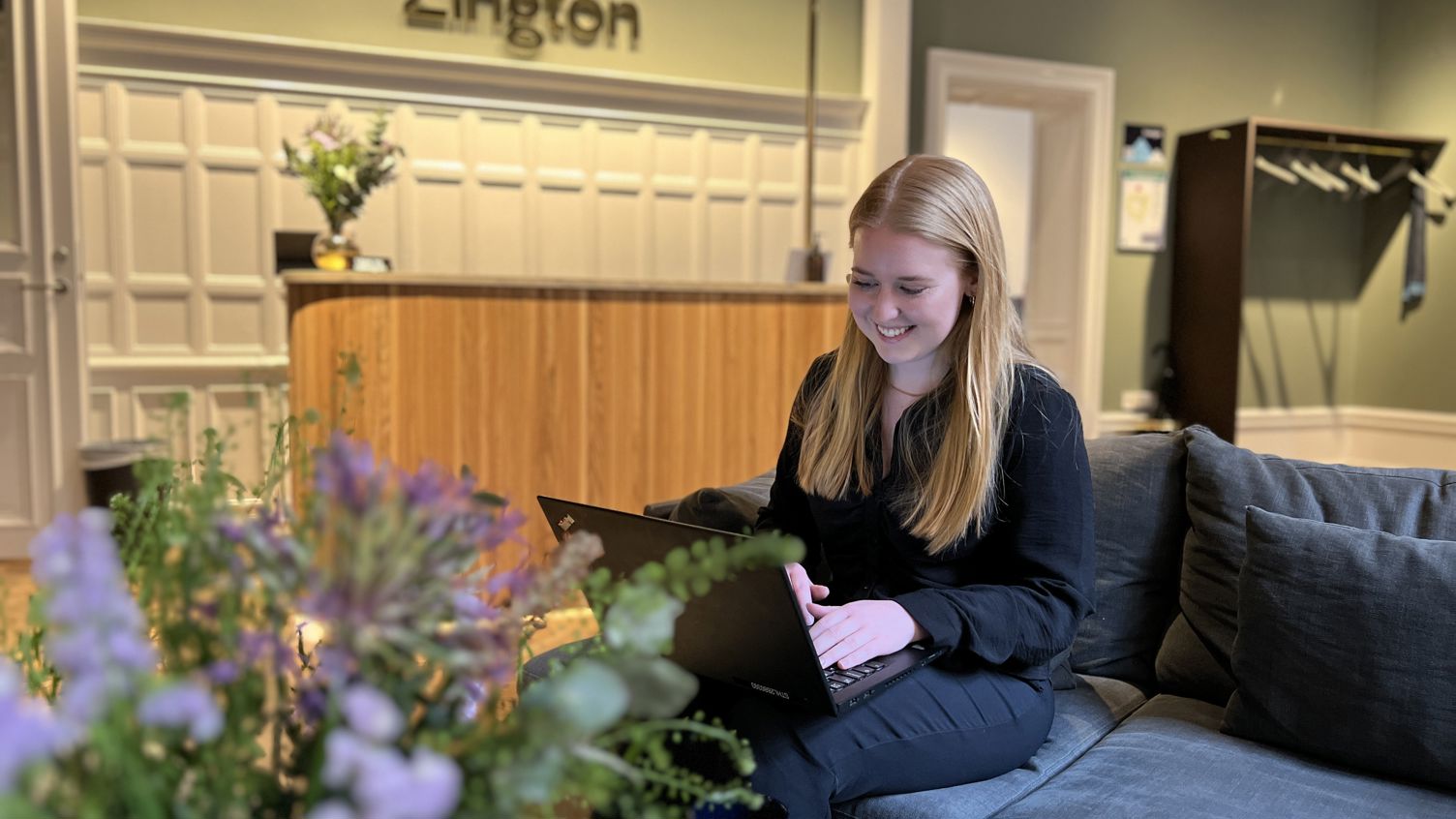 Zingtonmedarbetaren Melanie sitter i en soffa i Zingtons gröna lobby med en dator i famnen, hon ler.